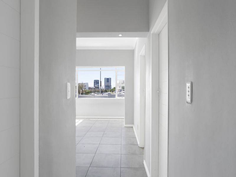 A bright, neutral-toned hallway featuring tiled flooring, recessed lighting, and clean finishes, leading towards an open living area with city views.