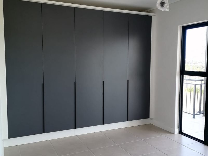 A bright, modern bedroom featuring sleek dark grey built-in cupboards, neutral floor tiles, and direct access to an outdoor balcony through a glass door.