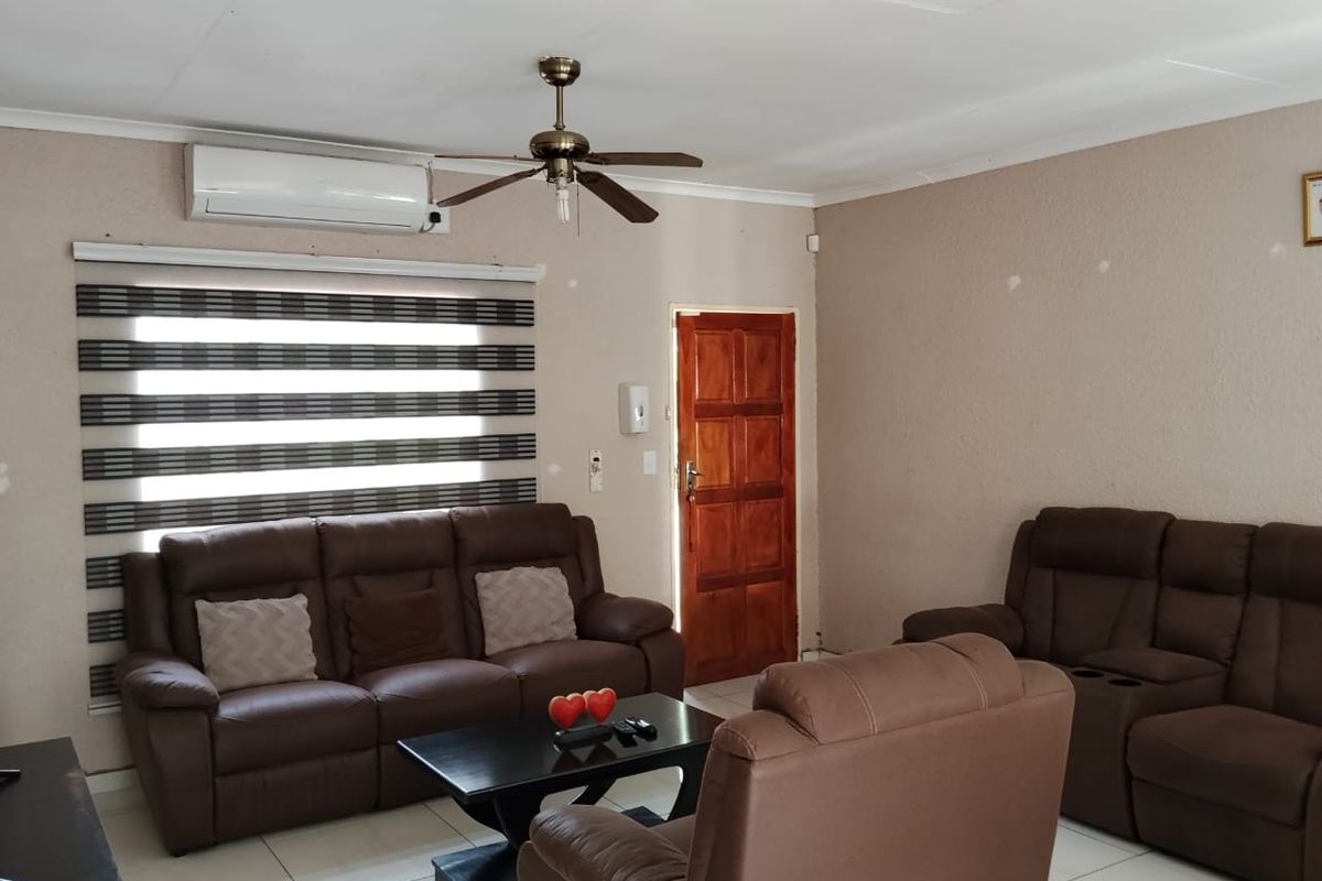 A living room featuring tiled flooring, a ceiling fan, air conditioning, and a wooden entrance door with modern roller blinds.
