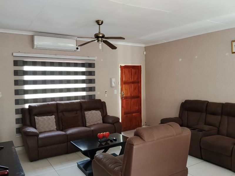 A living room featuring tiled flooring, a ceiling fan, air conditioning, and a wooden entrance door with modern roller blinds.