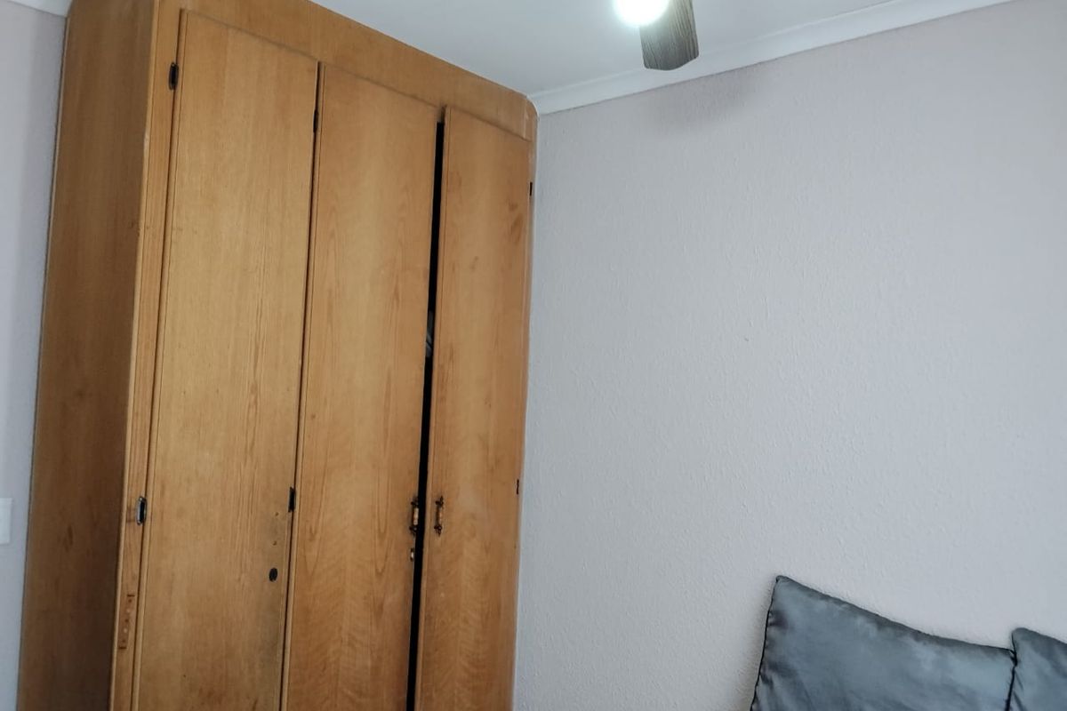 A bedroom featuring a large built-in wooden wardrobe and a ceiling fan, showing signs of wear and dated finishes.