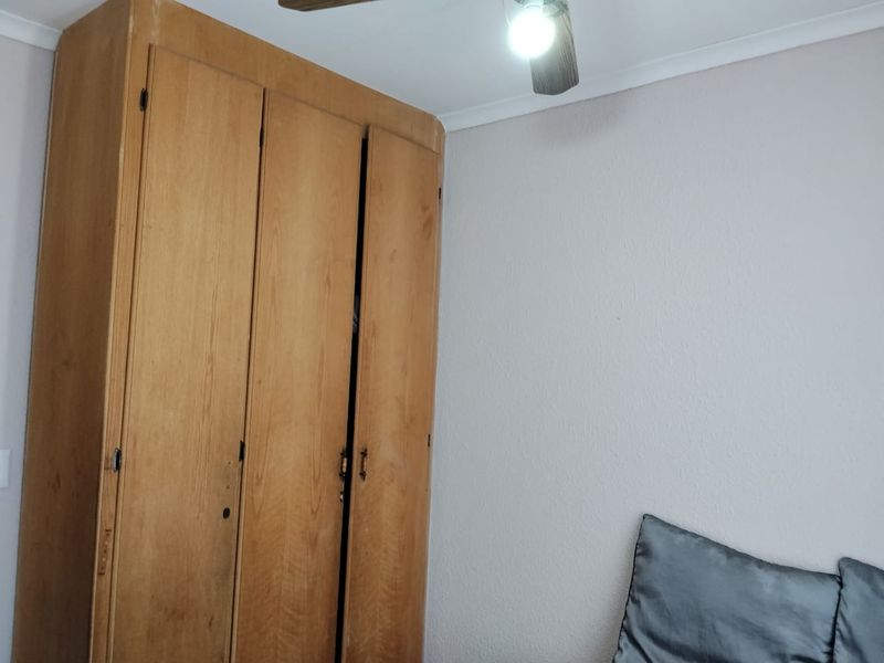 A bedroom featuring a large built-in wooden wardrobe and a ceiling fan, showing signs of wear and dated finishes.
