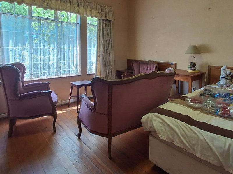 A bedroom featuring hardwood flooring and a large window, currently showing signs of wear on the wall surfaces.