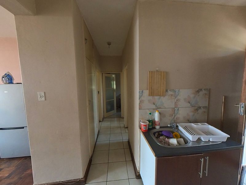 Open plan living area featuring tiled flooring, a compact kitchen sink unit with splashback, and access to a hallway leading to other rooms.