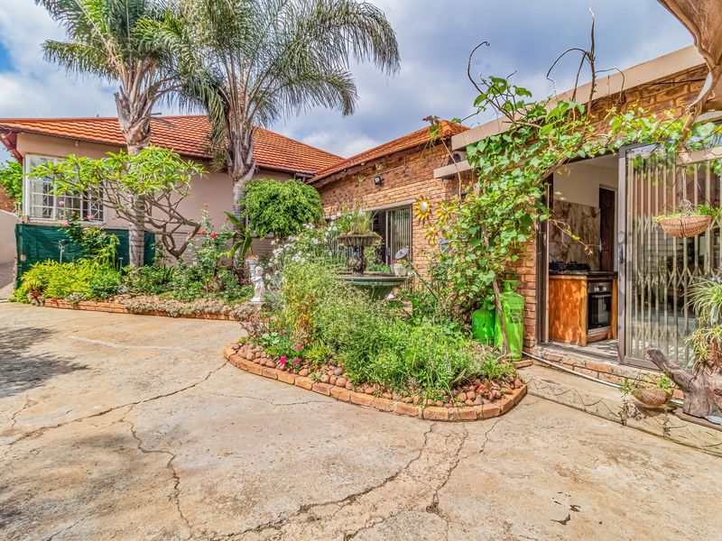 Rear exterior view featuring a brick facade, concrete patio area, established garden beds, and access to an indoor kitchen space via sliding doors.