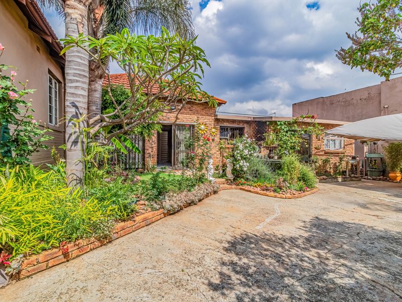 A rear exterior view of the house featuring a brick facade, tiled roof, and a concrete driveway surrounded by established garden beds.