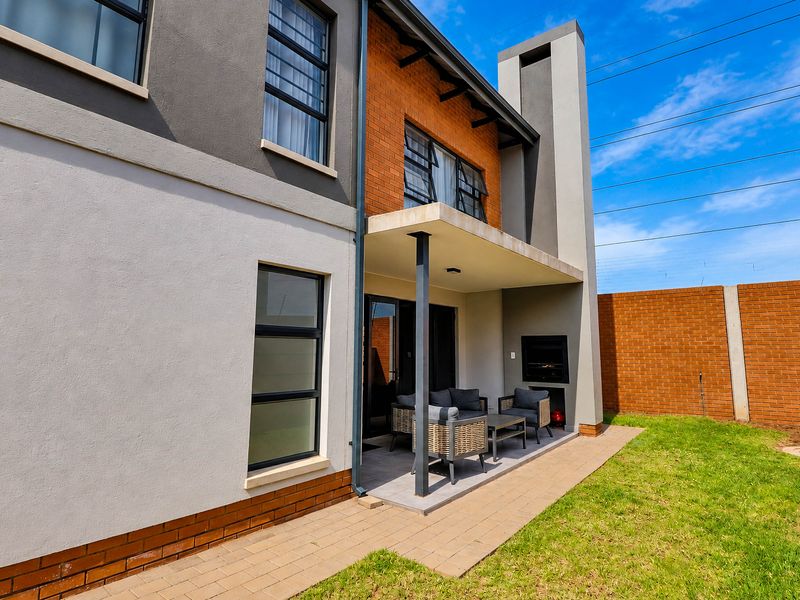 Rear view of the townhouse featuring a covered patio with a built-in braai, paved walkway, and a private garden area with brick boundary walls.