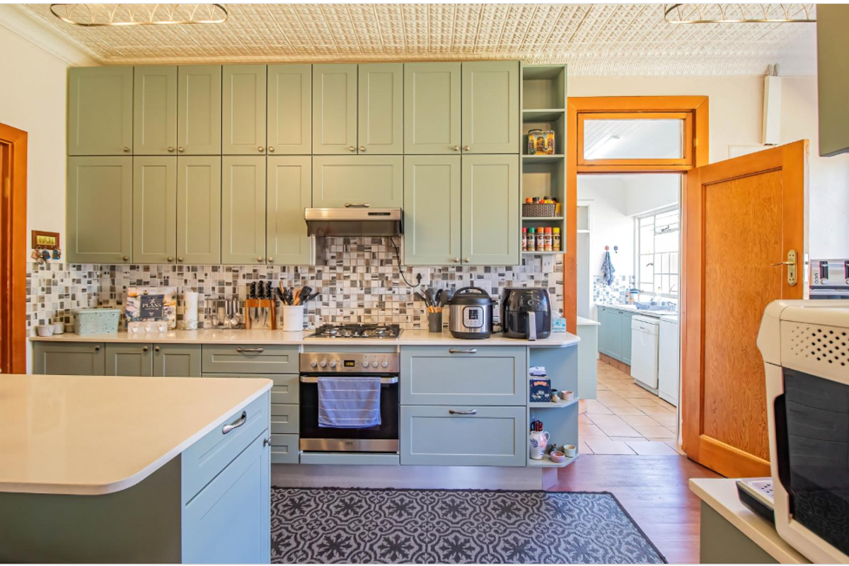 A well-maintained kitchen featuring sage green cabinetry, stone benchtops, a gas cooktop, and a distinctive pressed metal ceiling, with an adjacent scullery area.