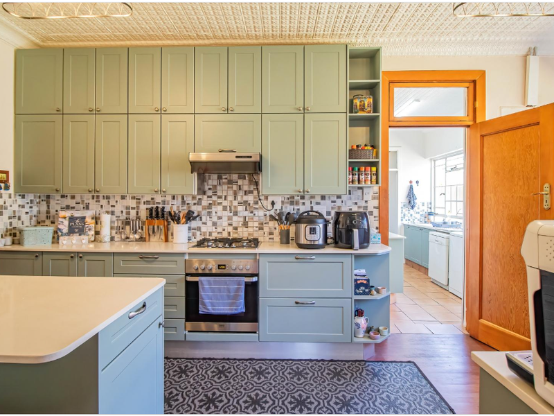 A well-maintained kitchen featuring sage green cabinetry, stone benchtops, a gas cooktop, and a distinctive pressed metal ceiling, with an adjacent scullery area.