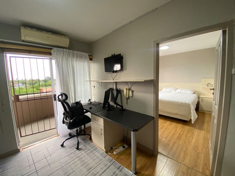 Living area featuring laminate flooring, a built-in desk, and a sliding door leading to a balcony, with an adjacent bedroom visible.