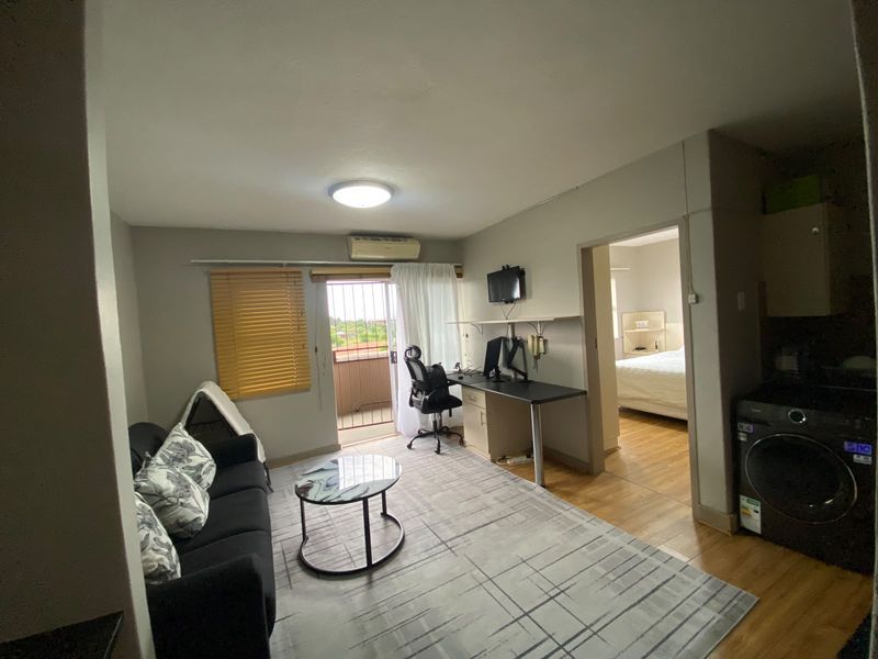 Open plan living area featuring laminate flooring, air conditioning, and direct access to a balcony, with an adjacent bedroom visible.
