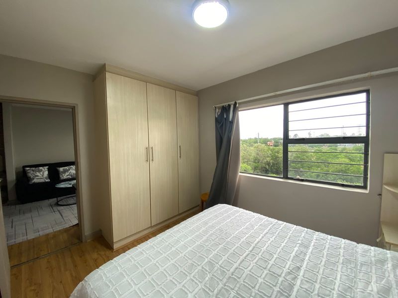 A well-maintained bedroom featuring built-in wardrobes, laminate flooring, and a window with security bars offering a pleasant green view.