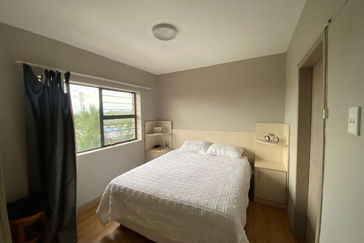 A functional bedroom featuring laminate flooring, built-in headboard with integrated bedside shelving, and a window with security bars.