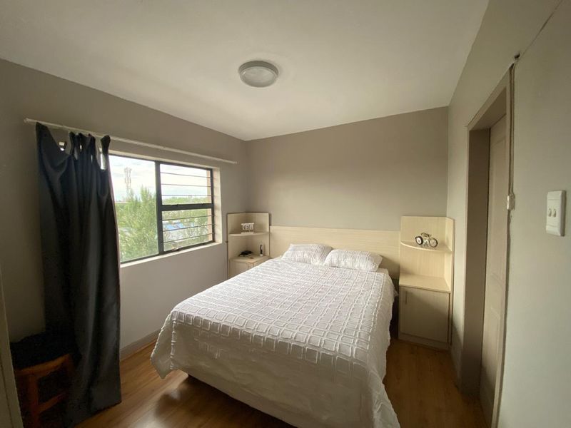 A functional bedroom featuring laminate flooring, built-in headboard with integrated bedside shelving, and a window with security bars.