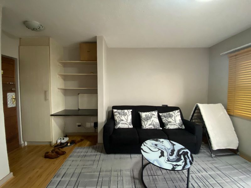 A living room featuring laminate flooring, built-in shelving with an integrated desk area, and window blinds.