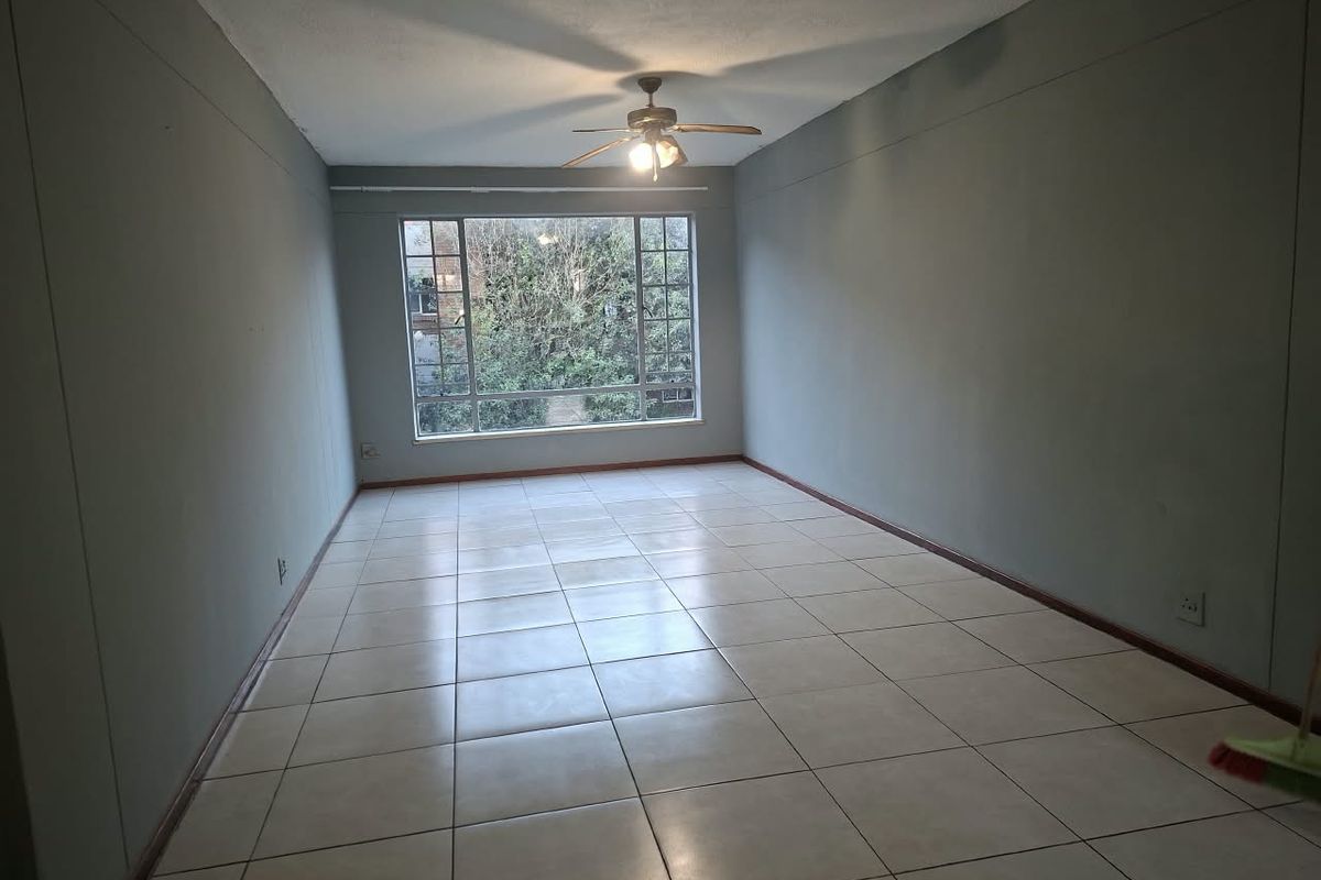 A spacious, empty living area featuring neutral tiled flooring, a ceiling fan, and a large window providing natural light and garden views.
