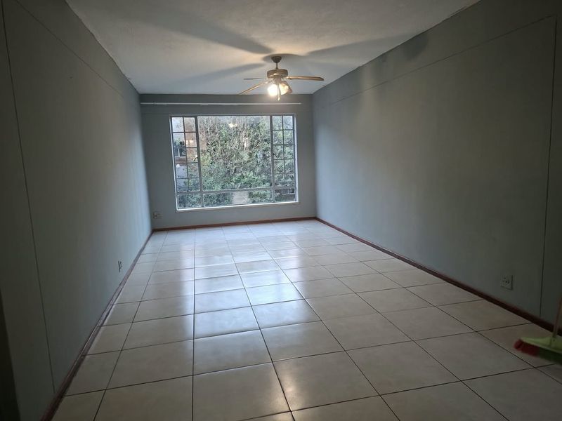 A spacious, empty living area featuring neutral tiled flooring, a ceiling fan, and a large window providing natural light and garden views.