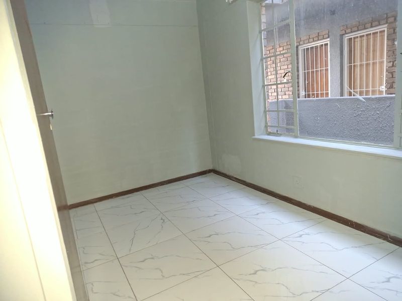 An empty bedroom featuring white marble-look floor tiles, steel window frames with security bars, and walls requiring final finishing and painting.