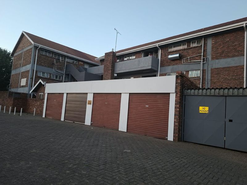 Exterior view of a brick apartment complex featuring multiple roller shutter garage doors, a paved driveway, and secure gated access.