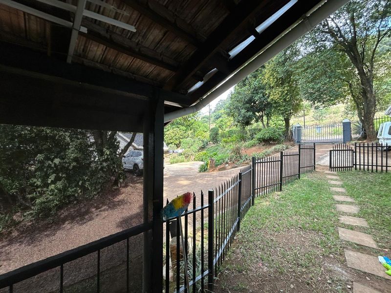 A covered patio area overlooking the garden and paved driveway, featuring a timber roof structure and secure metal perimeter fencing.