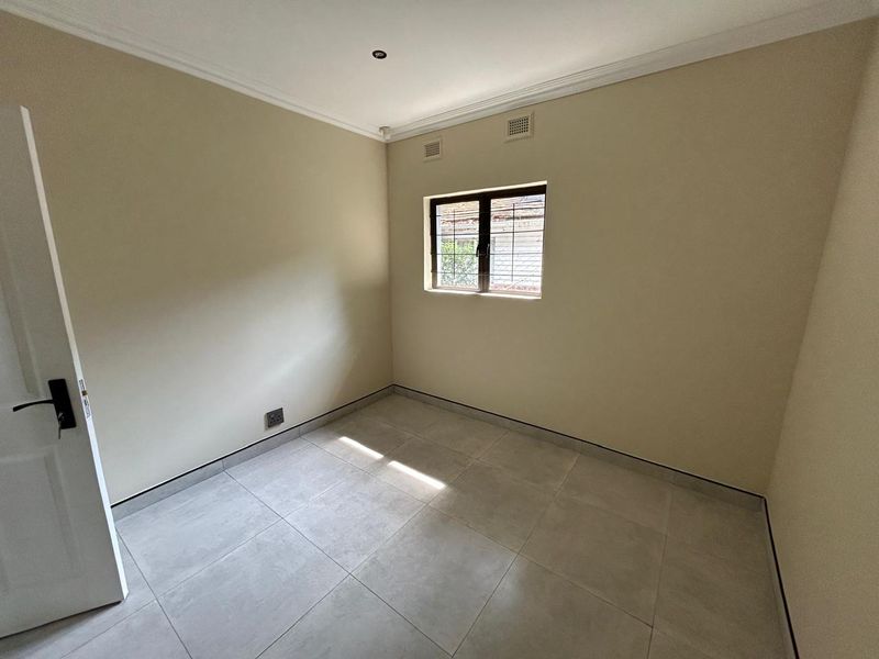 A clean, empty bedroom featuring neutral wall paint, modern grey floor tiles, coving, and a window fitted with security bars.