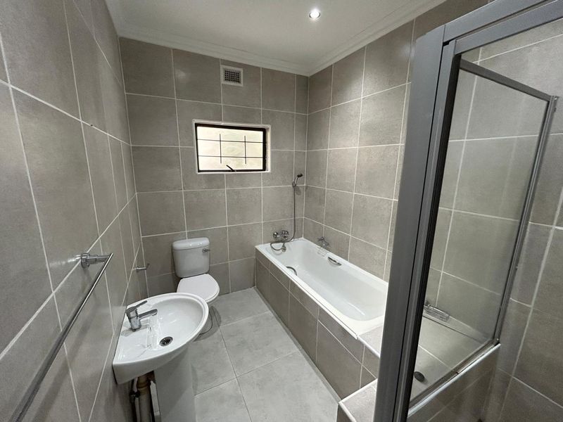 A clean, modern bathroom featuring neutral floor-to-ceiling tiling, a pedestal basin, a toilet, and a combined bathtub and shower enclosure.