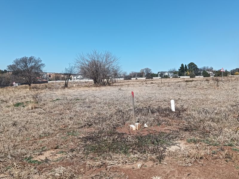 A vacant residential plot of land featuring natural grass cover and visible boundary survey markers under a clear blue sky.