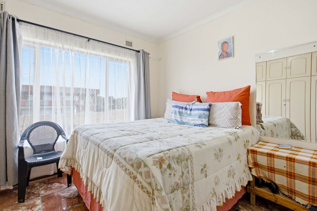 A bedroom featuring built-in cupboards, tiled flooring, and a large window with a security gate, offering natural light.