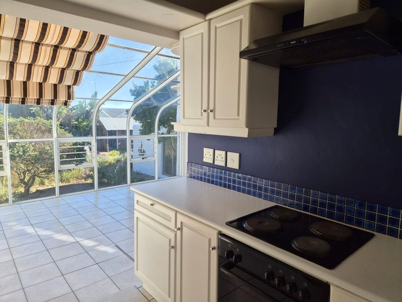A bright kitchen featuring built-in cabinetry, an electric hob, and an extractor fan, seamlessly connected to a sun-filled glass conservatory area.
