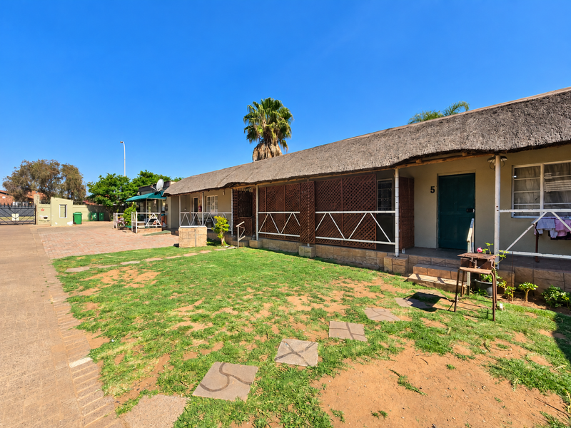 Rear view of a retirement village unit featuring a traditional thatch roof, paved access, and private entrance with lattice screening.