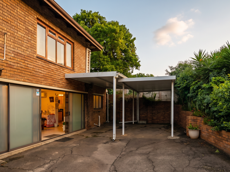Rear exterior view featuring a brick facade, sliding glass doors, a covered carport area, and a paved driveway with established garden borders.