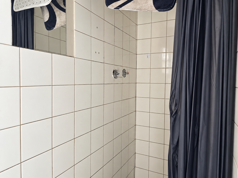 A functional bathroom shower area featuring white ceramic wall tiles, a wall-mounted mixer, and a built-in mirror, currently requiring minor maintenance and fixture updates.