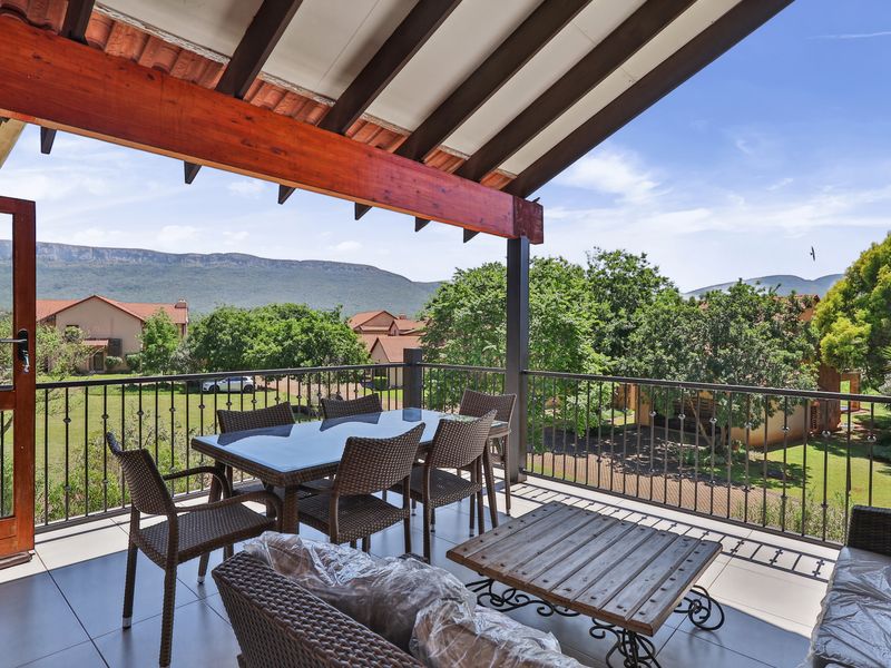 A spacious covered balcony featuring tiled flooring, exposed timber rafters, and a wrought iron balustrade, offering scenic views of the surrounding landscape.
