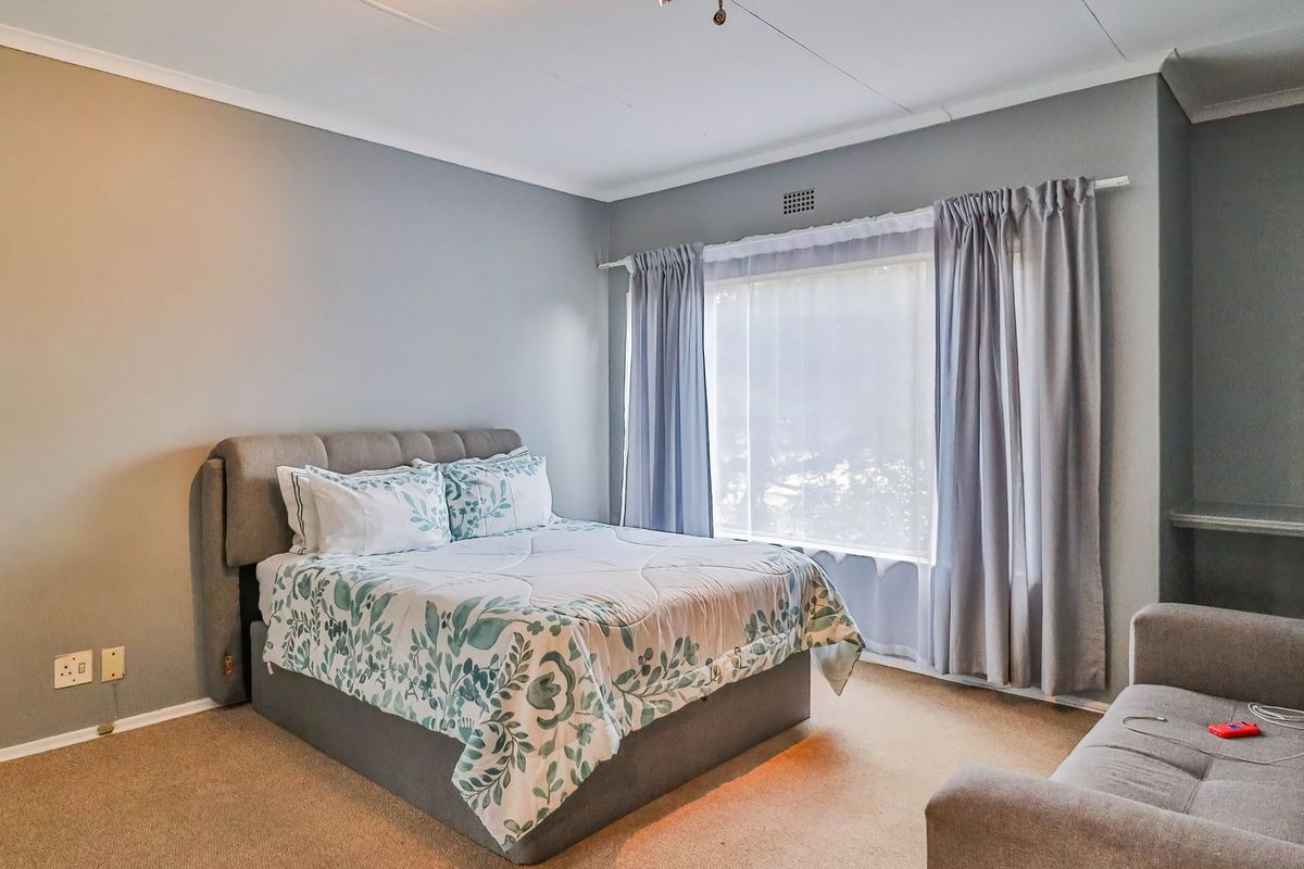 A well-lit bedroom featuring neutral wall tones, carpeted flooring, and a large window with installed curtain tracks.