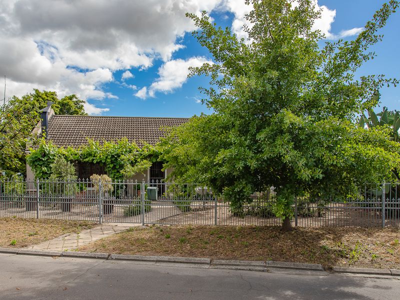 Front view of a single-story house featuring a tiled roof, chimney, and metal perimeter fencing with a paved walkway leading to the entrance.