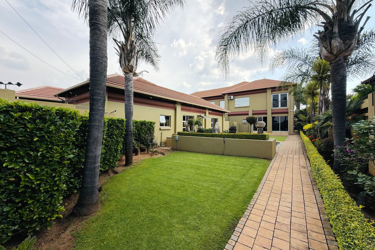 A well-maintained rear garden featuring a paved walkway, manicured lawn, mature palm trees, and established hedging alongside the house exterior.