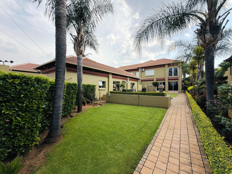 A well-maintained rear garden featuring a paved walkway, manicured lawn, mature palm trees, and established hedging alongside the house exterior.