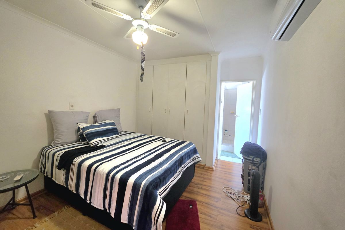 A functional bedroom featuring laminate flooring, built-in cupboards, a ceiling fan, and a wall-mounted air conditioning unit, with direct access to an en-suite bathroom.