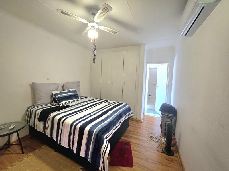 A functional bedroom featuring laminate flooring, built-in cupboards, a ceiling fan, and a wall-mounted air conditioning unit, with direct access to an en-suite bathroom.