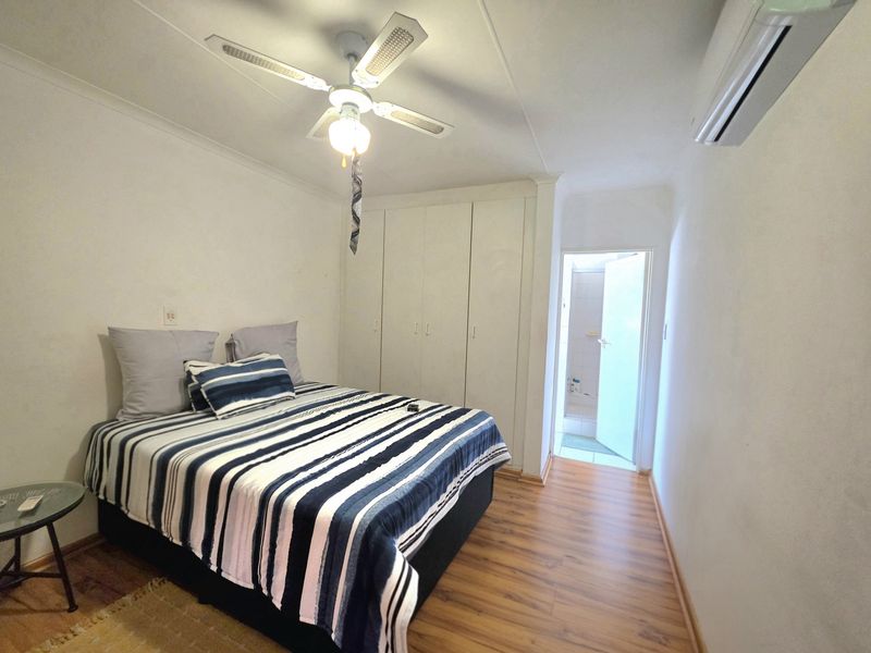 A functional bedroom featuring built-in cupboards, laminate flooring, a ceiling fan, and a wall-mounted air conditioning unit, with an adjoining bathroom visible.