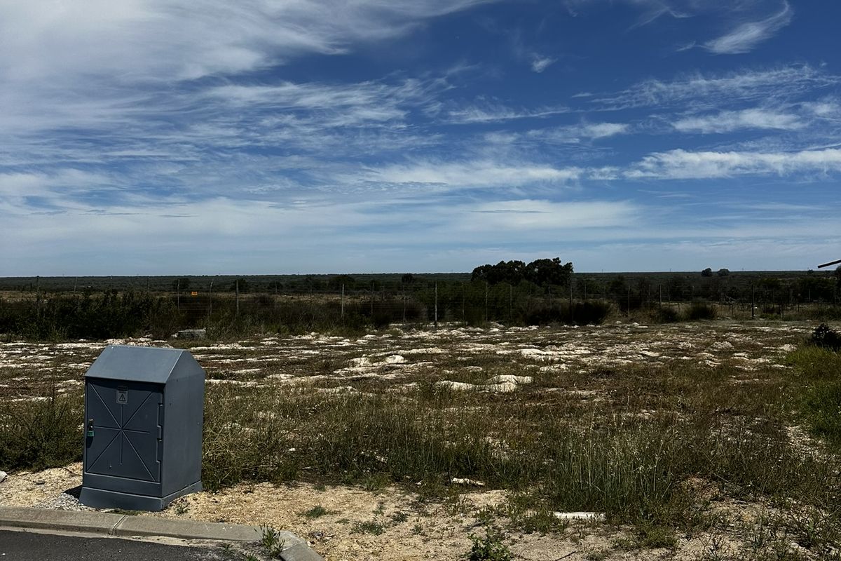 A vacant residential plot featuring an asphalt road frontage, concrete curbing, and an installed electrical utility box, ready for development.