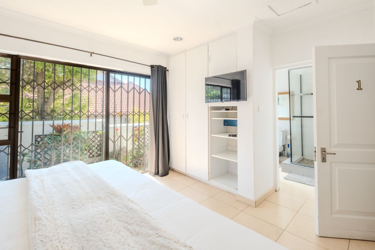 A bright bedroom featuring tiled floors, built-in cupboards, and a sliding door with security gate, leading to an en-suite bathroom.