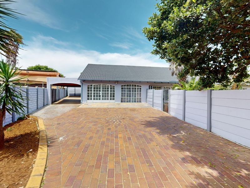 Front view of the property featuring a spacious brick-paved driveway, a carport, and a secure boundary wall with arched glass entrance doors.