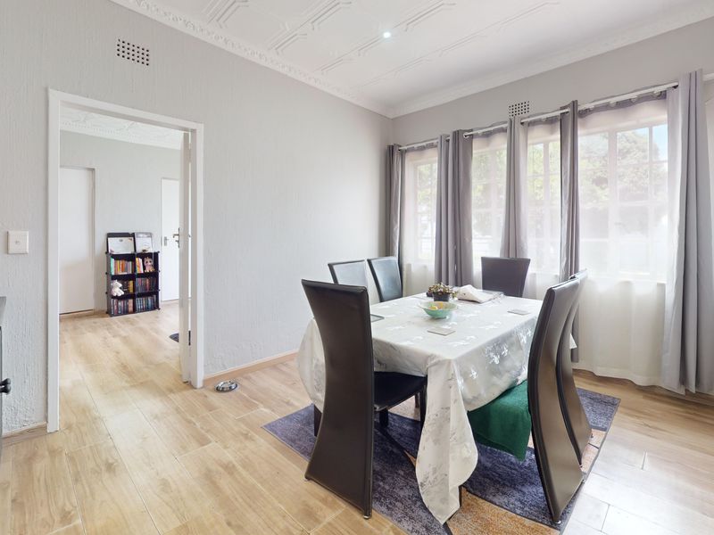 A bright dining room featuring light laminate flooring, textured walls, and decorative ceiling cornicing, illuminated by large multi-pane windows.