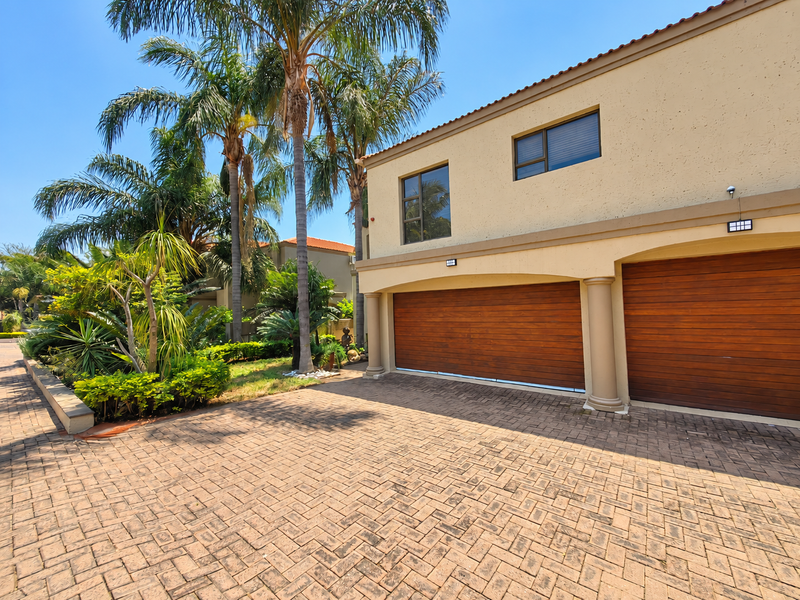 Front elevation of a townhouse featuring a double garage with wooden doors, paved driveway, and established garden landscaping.