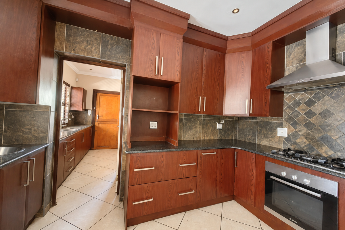 A spacious kitchen featuring dark wood cabinetry, granite countertops, built-in oven, gas hob, and a tiled backsplash, with an adjacent scullery area.