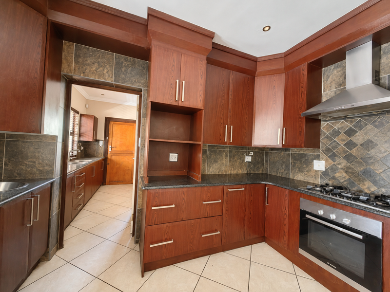 A spacious kitchen featuring dark wood cabinetry, granite countertops, built-in oven, gas hob, and a tiled backsplash, with an adjacent scullery area.
