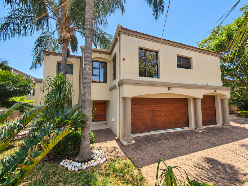 A well-maintained double-story townhouse featuring a paved driveway, double wooden garage doors, and decorative architectural columns under a tiled roof.