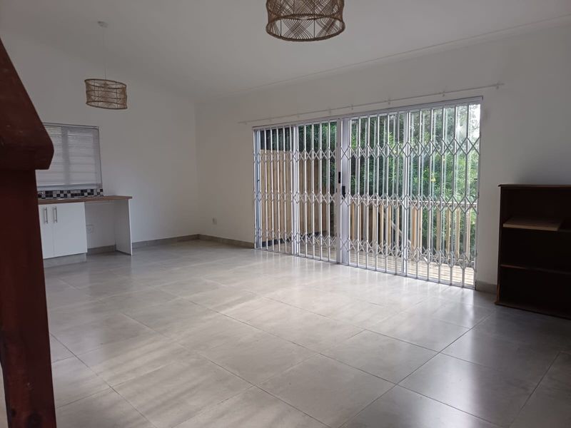Spacious open-plan living area featuring large format floor tiles, a secure sliding door leading to the exterior, and a compact kitchen workspace.