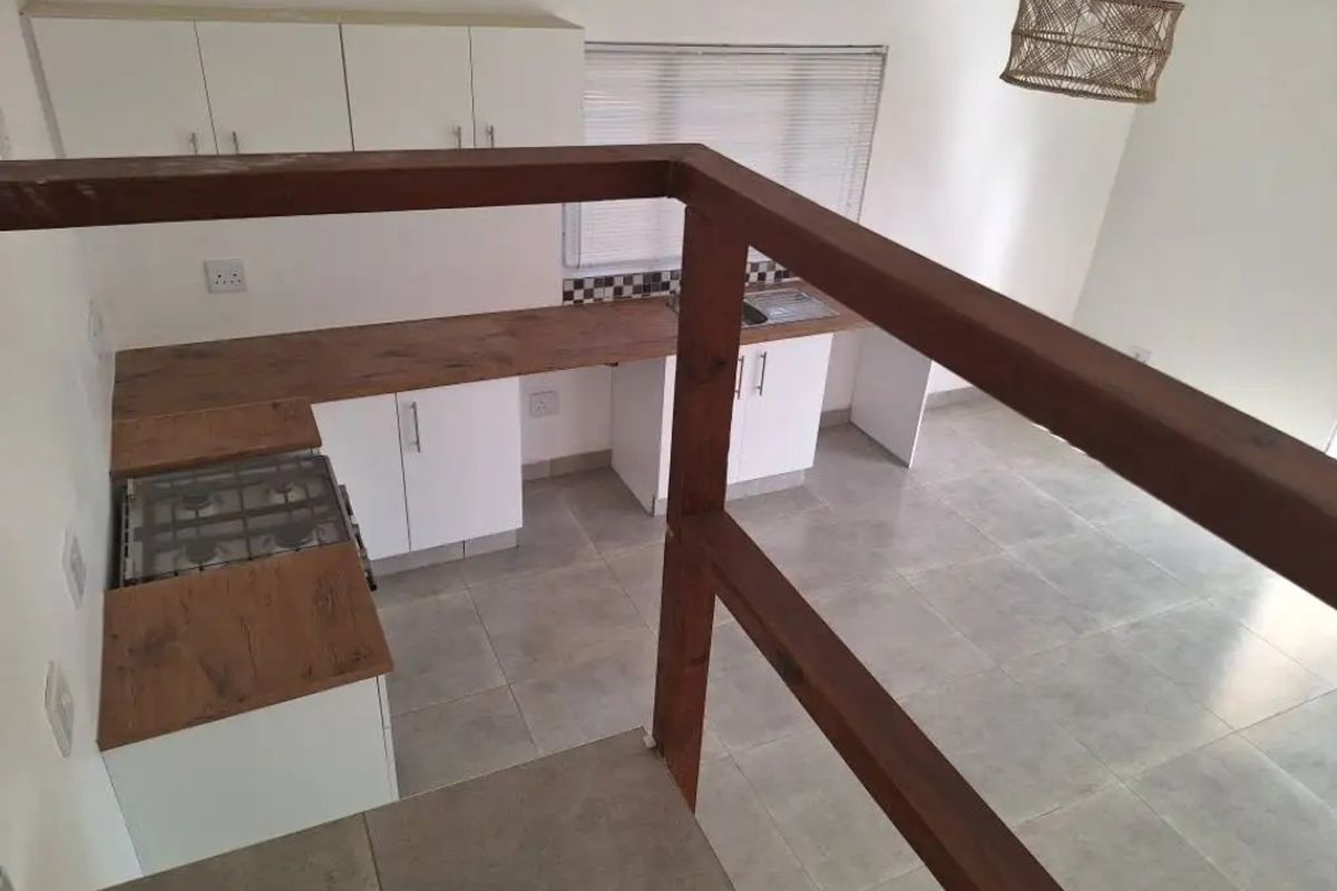 A clean, modern kitchen featuring white cabinetry, wood-look countertops, a gas hob, and tiled flooring, viewed from an elevated position behind a timber balustrade.